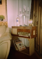 A cozy corner featuring a vintage wooden radio on a simple wooden stand, adorned with a silver candelabra, a globe, and a clear bottle. Beside the radio, there is a collection of vinyl records leaning against the stand. The setting includes a beige leather sofa and soft white curtains, with logs stacked against the wall as part of the decor. A small potted plant adds a touch of greenery to the rustic atmosphere.