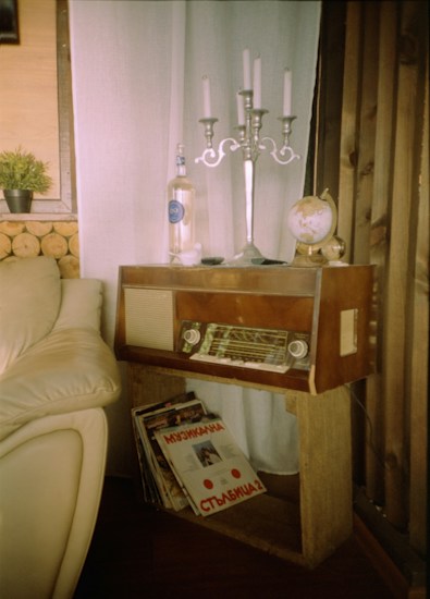 A cozy corner featuring a vintage wooden radio on a simple wooden stand, adorned with a silver candelabra, a globe, and a clear bottle. Beside the radio, there is a collection of vinyl records leaning against the stand. The setting includes a beige leather sofa and soft white curtains, with logs stacked against the wall as part of the decor. A small potted plant adds a touch of greenery to the rustic atmosphere.