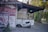 A small scooter is parked on a stone path next to a building with tiled roofing. The building has a sign with various food items and the words 'fast food' prominently displayed. There are trees and greenery surrounding the area, contributing to a tranquil setting.