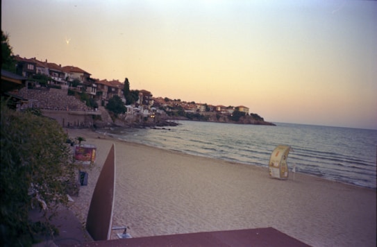 A tranquil beach scene at dusk with a gentle shoreline and a small village with red-roofed buildings nestled along the coast. The sky is a gradient of soft pastel hues, and distant hills are visible. There is minimal human presence, with a single structure that resembles a small kiosk located on the sandy shore.