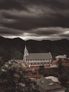 A large church with a prominent white facade and a tall steeple is situated on a hillside surrounded by lush green forests. Overhead, heavy and dark clouds create a dramatic and moody atmosphere. Nearby, several smaller buildings are visible, contributing to a rustic and serene landscape.