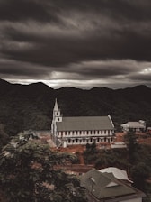 A large church with a prominent white facade and a tall steeple is situated on a hillside surrounded by lush green forests. Overhead, heavy and dark clouds create a dramatic and moody atmosphere. Nearby, several smaller buildings are visible, contributing to a rustic and serene landscape.