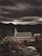 A large church with a prominent white facade and a tall steeple is situated on a hillside surrounded by lush green forests. Overhead, heavy and dark clouds create a dramatic and moody atmosphere. Nearby, several smaller buildings are visible, contributing to a rustic and serene landscape.