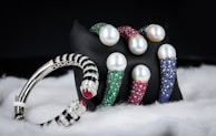 A collection of luxury bracelets and bangles featuring large pearls and encrusted with multicolored gemstones is displayed against a dark background. The bracelets rest on a black stand and are surrounded by a soft, white, fabric-like material. The gemstones include green, red, and blue hues, while the metal appears to be silver or platinum.