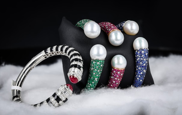 A collection of luxury bracelets and bangles featuring large pearls and encrusted with multicolored gemstones is displayed against a dark background. The bracelets rest on a black stand and are surrounded by a soft, white, fabric-like material. The gemstones include green, red, and blue hues, while the metal appears to be silver or platinum.