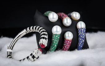 A collection of luxury bracelets and bangles featuring large pearls and encrusted with multicolored gemstones is displayed against a dark background. The bracelets rest on a black stand and are surrounded by a soft, white, fabric-like material. The gemstones include green, red, and blue hues, while the metal appears to be silver or platinum.