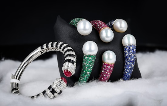 A collection of luxury bracelets and bangles featuring large pearls and encrusted with multicolored gemstones is displayed against a dark background. The bracelets rest on a black stand and are surrounded by a soft, white, fabric-like material. The gemstones include green, red, and blue hues, while the metal appears to be silver or platinum.