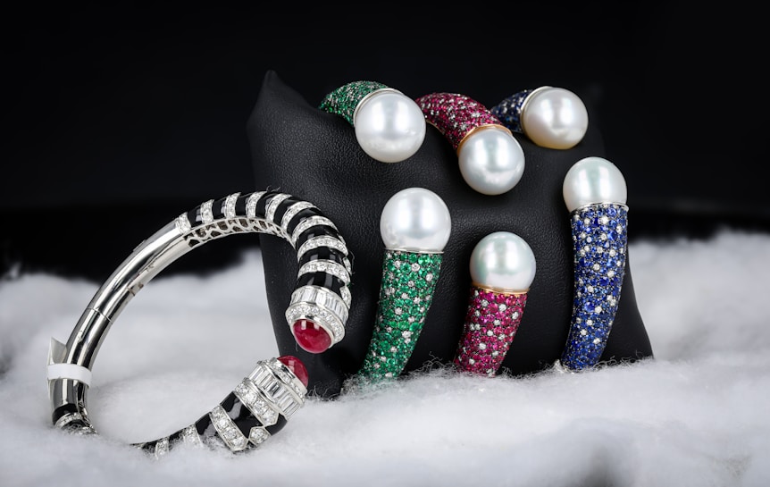 A collection of luxury bracelets and bangles featuring large pearls and encrusted with multicolored gemstones is displayed against a dark background. The bracelets rest on a black stand and are surrounded by a soft, white, fabric-like material. The gemstones include green, red, and blue hues, while the metal appears to be silver or platinum.