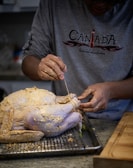 A person wearing a gray long-sleeve shirt is preparing a raw, seasoned turkey on a wire rack placed over a baking tray. The person is using a string or twine to truss the turkey, with hands covered in seasoning.