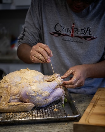 A person wearing a gray long-sleeve shirt is preparing a raw, seasoned turkey on a wire rack placed over a baking tray. The person is using a string or twine to truss the turkey, with hands covered in seasoning.