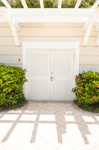 A white double door with louvered panels is set into a light beige building wall. Two lush green bushes with red flowers flank the doorway. The ground is covered with light-colored tiles, and geometric shadows from a pergola overhead cross the floor. Above the door, part of a trellis is visible, and hints of greenery are seen at the top.