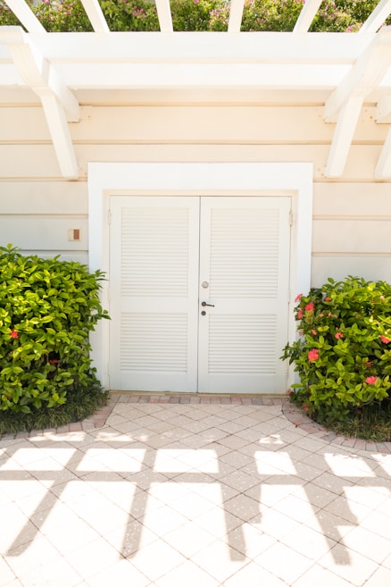 A white double door with louvered panels is set into a light beige building wall. Two lush green bushes with red flowers flank the doorway. The ground is covered with light-colored tiles, and geometric shadows from a pergola overhead cross the floor. Above the door, part of a trellis is visible, and hints of greenery are seen at the top.