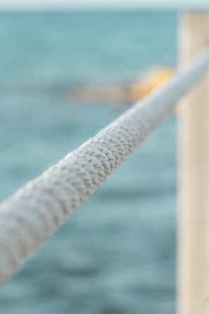 A close-up view of a taut, textured rope with a blurred background of the sea. The rope spans diagonally across the image, highlighting its intricate weave.