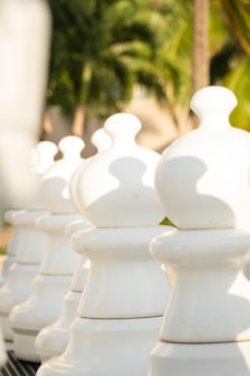 Large white chess pawn pieces are aligned on an outdoor board, with a blurred background of tropical palm trees and greenery. The sunlight casts shadows on the pieces, creating a serene and leisurely atmosphere.