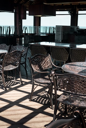An outdoor dining area features intricate metal chairs and tables under a wooden canopy. Sunlight casts geometric shadows on the wooden floor, while the sea is visible in the background. Chairs are neatly stacked in the back.