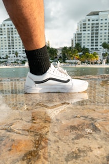A leg wearing a black sock and a white sneaker with a black stripe is positioned on a wet stone surface. The background features a waterfront scene with tall white buildings, palm trees, and a partly cloudy sky.