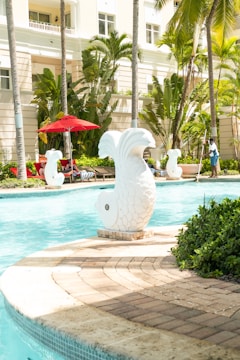 A tranquil poolside scene with decorative white fish statues surrounding a clear blue swimming pool. Lush green palm trees and plants provide a tropical background. Sun loungers with red umbrellas are arranged near the water, creating a relaxing atmosphere. A person is seen standing with a net, possibly cleaning the pool.