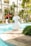 A tranquil poolside scene with decorative white fish statues surrounding a clear blue swimming pool. Lush green palm trees and plants provide a tropical background. Sun loungers with red umbrellas are arranged near the water, creating a relaxing atmosphere. A person is seen standing with a net, possibly cleaning the pool.