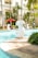 A tranquil poolside scene with decorative white fish statues surrounding a clear blue swimming pool. Lush green palm trees and plants provide a tropical background. Sun loungers with red umbrellas are arranged near the water, creating a relaxing atmosphere. A person is seen standing with a net, possibly cleaning the pool.
