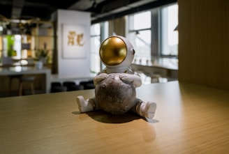 A small, futuristic figurine with a gold spherical helmet sits on a wooden surface, holding what appears to be a small moon-like object. The background is a modern room with soft lighting, blurred plants, and abstract art on the wall.