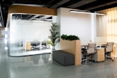 A modern office space featuring a glass-walled conference room with chairs and a desk visible inside. Adjacent are open workstations with ergonomic chairs and a gray cushioned bench. The decor includes potted plants, providing a touch of greenery. The room is well-lit with a linear overhead light and ambient sunlight coming through window blinds.