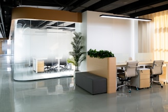 A modern office space featuring a glass-walled conference room with chairs and a desk visible inside. Adjacent are open workstations with ergonomic chairs and a gray cushioned bench. The decor includes potted plants, providing a touch of greenery. The room is well-lit with a linear overhead light and ambient sunlight coming through window blinds.