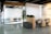 A modern office space featuring a glass-walled conference room with chairs and a desk visible inside. Adjacent are open workstations with ergonomic chairs and a gray cushioned bench. The decor includes potted plants, providing a touch of greenery. The room is well-lit with a linear overhead light and ambient sunlight coming through window blinds.