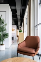 A modern interior space featuring a brown leather chair in the foreground with a light wood table. The room has a sleek design, with a white wall and green potted plant on the left side. Natural light pours in from large windows, partially covered by beige curtains. In the background, there's a long narrow space with wooden furniture and pendant lights, leading to a distant focal point with artwork on the wall.