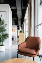 A modern interior space featuring a brown leather chair in the foreground with a light wood table. The room has a sleek design, with a white wall and green potted plant on the left side. Natural light pours in from large windows, partially covered by beige curtains. In the background, there's a long narrow space with wooden furniture and pendant lights, leading to a distant focal point with artwork on the wall.