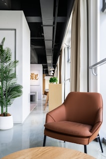 A modern interior space featuring a brown leather chair in the foreground with a light wood table. The room has a sleek design, with a white wall and green potted plant on the left side. Natural light pours in from large windows, partially covered by beige curtains. In the background, there's a long narrow space with wooden furniture and pendant lights, leading to a distant focal point with artwork on the wall.