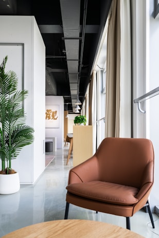 A modern interior space featuring a brown leather chair in the foreground with a light wood table. The room has a sleek design, with a white wall and green potted plant on the left side. Natural light pours in from large windows, partially covered by beige curtains. In the background, there's a long narrow space with wooden furniture and pendant lights, leading to a distant focal point with artwork on the wall.
