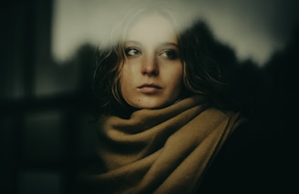 A person appears to be gazing thoughtfully through a blurred window, wrapped in a beige scarf. The lighting is soft and subdued, creating a contemplative and introspective atmosphere.