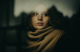 A person appears to be gazing thoughtfully through a blurred window, wrapped in a beige scarf. The lighting is soft and subdued, creating a contemplative and introspective atmosphere.