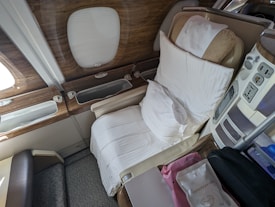 A luxurious airplane seat designed for comfort, upholstered in beige leather with a plush pillow and sheet covering. A high-quality wooden panel is near the window, creating a serene atmosphere. Nearby are convenient features such as power outlets and a control panel, along with personal items including a soft storage bag.