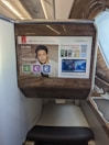 The photo features a screen in an airplane, displaying flight information from Emirates airline for flight EK225 from Dubai to San Francisco in seat 14K. The screen interface offers options like information, communication, and entertainment. Advertisements and travel details, including a map and promotional offers, are visible. The surroundings exhibit luxury finishes with wood paneling.
