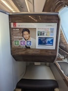 The photo features a screen in an airplane, displaying flight information from Emirates airline for flight EK225 from Dubai to San Francisco in seat 14K. The screen interface offers options like information, communication, and entertainment. Advertisements and travel details, including a map and promotional offers, are visible. The surroundings exhibit luxury finishes with wood paneling.