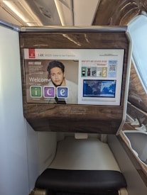 The photo features a screen in an airplane, displaying flight information from Emirates airline for flight EK225 from Dubai to San Francisco in seat 14K. The screen interface offers options like information, communication, and entertainment. Advertisements and travel details, including a map and promotional offers, are visible. The surroundings exhibit luxury finishes with wood paneling.