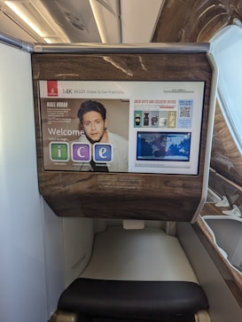 The photo features a screen in an airplane, displaying flight information from Emirates airline for flight EK225 from Dubai to San Francisco in seat 14K. The screen interface offers options like information, communication, and entertainment. Advertisements and travel details, including a map and promotional offers, are visible. The surroundings exhibit luxury finishes with wood paneling.