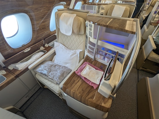A luxurious airplane cabin with a plush reclining seat featuring a quilted design. The seat is complemented by a pillow and a packaged blanket, creating a comfortable environment. Adjacent to the seat is a private mini-bar stocked with bottled water and other refreshments. The interior is accentuated with rich wooden paneling and modern technological amenities for convenience and relaxation.
