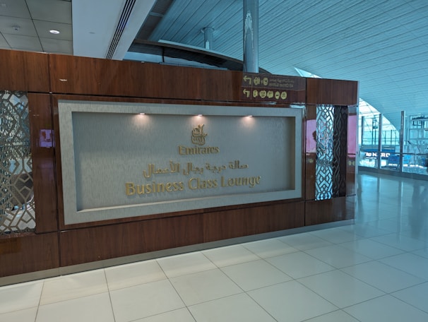 A signboard for the Business Class Lounge of Emirates airline is mounted on a wooden wall. The text 'Emirates Business Class Lounge' is written in both English and Arabic in gold lettering. The floor is white tiled, and there are some decorative elements on the side panels of the wall. The background shows part of an airport terminal with glass windows and visible signage.