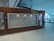 A signboard for the Business Class Lounge of Emirates airline is mounted on a wooden wall. The text 'Emirates Business Class Lounge' is written in both English and Arabic in gold lettering. The floor is white tiled, and there are some decorative elements on the side panels of the wall. The background shows part of an airport terminal with glass windows and visible signage.
