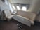 The image depicts a luxurious seating area inside an airplane. The space features a cream-colored sofa and a matching seat aligned along the wall, underneath three windows. There is a wooden table with a metallic base positioned in front of the seating. The area appears sophisticated with beige curtains framing the windows and a small lamp on a side table.