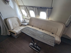 The image depicts a luxurious seating area inside an airplane. The space features a cream-colored sofa and a matching seat aligned along the wall, underneath three windows. There is a wooden table with a metallic base positioned in front of the seating. The area appears sophisticated with beige curtains framing the windows and a small lamp on a side table.