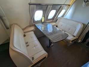 A luxurious seating area inside a vehicle, featuring two cream-colored leather couches with seatbelts, placed opposite each other across a polished wooden table. The setting is well-lit, with windows covered by elegant curtains, suggesting it's part of an airplane.