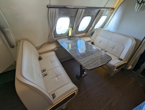 A luxurious seating area inside a vehicle, featuring two cream-colored leather couches with seatbelts, placed opposite each other across a polished wooden table. The setting is well-lit, with windows covered by elegant curtains, suggesting it's part of an airplane.