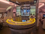 Luxurious airplane bar with a sleek, curved counter illuminated by warm, ambient lighting. The shelves are stocked with various bottles of liquor, and glasses are neatly arranged. Small plates of food are displayed on the counter. Comfortable seating surrounds the bar area, while elegant decor adds a sophisticated touch.
