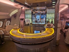 Luxurious airplane bar with a sleek, curved counter illuminated by warm, ambient lighting. The shelves are stocked with various bottles of liquor, and glasses are neatly arranged. Small plates of food are displayed on the counter. Comfortable seating surrounds the bar area, while elegant decor adds a sophisticated touch.