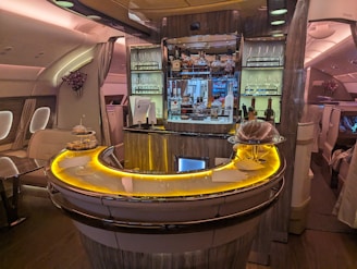 Luxurious airplane bar with a sleek, curved counter illuminated by warm, ambient lighting. The shelves are stocked with various bottles of liquor, and glasses are neatly arranged. Small plates of food are displayed on the counter. Comfortable seating surrounds the bar area, while elegant decor adds a sophisticated touch.