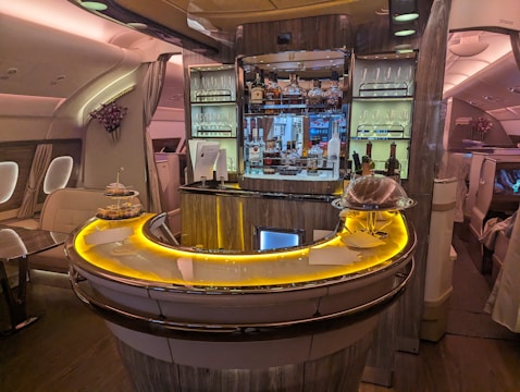 Luxurious airplane bar with a sleek, curved counter illuminated by warm, ambient lighting. The shelves are stocked with various bottles of liquor, and glasses are neatly arranged. Small plates of food are displayed on the counter. Comfortable seating surrounds the bar area, while elegant decor adds a sophisticated touch.