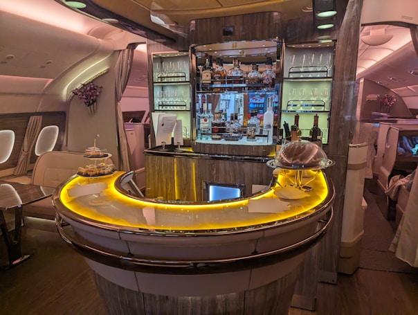 Luxurious airplane bar with a sleek, curved counter illuminated by warm, ambient lighting. The shelves are stocked with various bottles of liquor, and glasses are neatly arranged. Small plates of food are displayed on the counter. Comfortable seating surrounds the bar area, while elegant decor adds a sophisticated touch.